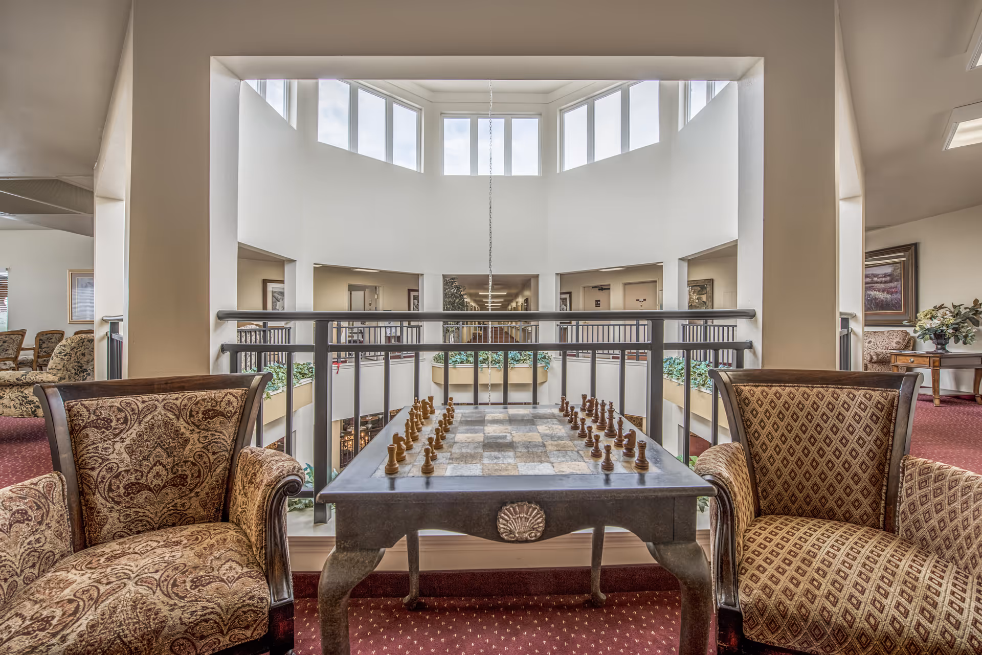 Upstairs seating area with a chess table between two upholstered chairs overlooking an open central atrium and high windows.
