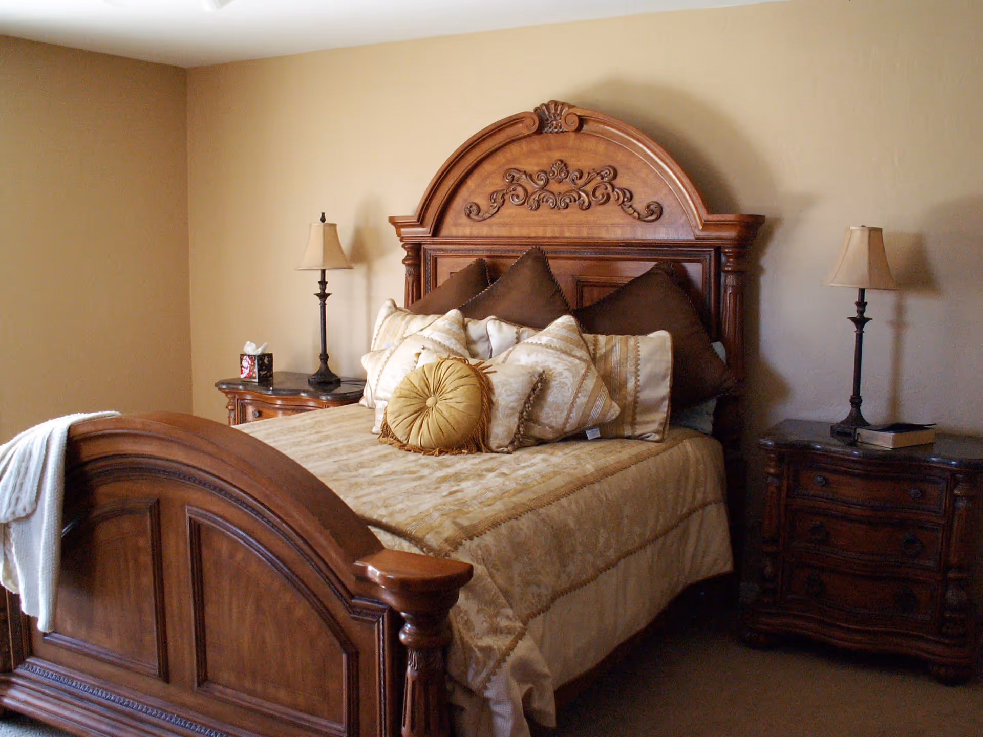 A bedroom with a large wooden bed featuring an ornate headboard and footboard. The bed is made with a beige and gold comforter and multiple decorative pillows in shades of brown and cream. There are two matching wooden nightstands on either side of the bed, each with a lamp and some items on top. The walls are painted a light beige color.