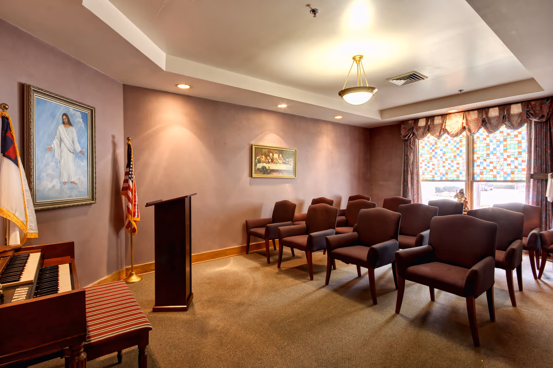 Small chapel-style meeting room with rows of upholstered chairs facing a wooden podium, an organ, flags, and religious artwork by patterned windows.