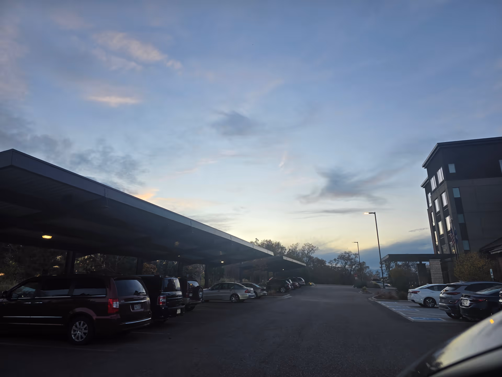 Parking lot with covered carports and multiple parked cars beside a multi-story building under a twilight sky.