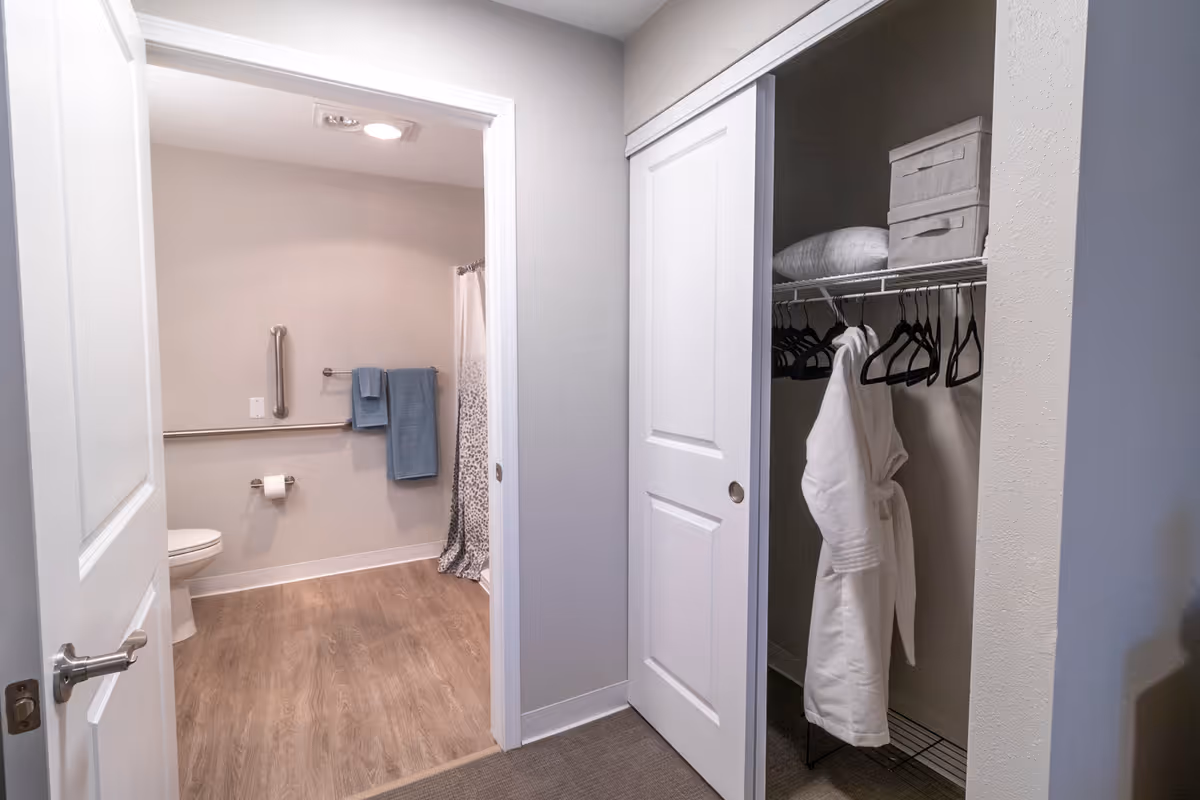 View of a bathroom with an open door showing a toilet, grab bars, a towel rack with blue towels, and a shower curtain. Adjacent to the bathroom is a closet with a white sliding door, containing a white bathrobe, hangers, a pillow, and storage boxes on a shelf.