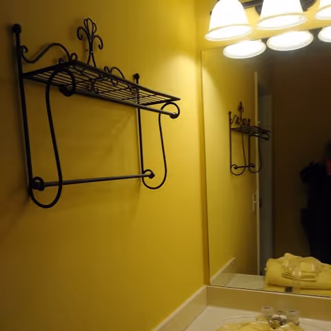 A bathroom interior with yellow walls, a metal wall-mounted towel rack, a large mirror reflecting the towel rack, a countertop with folded yellow towels, and overhead lights.