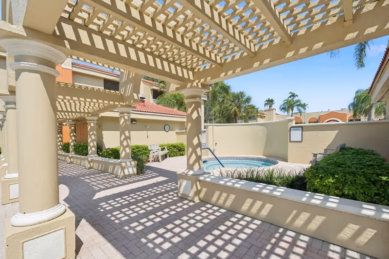 Outdoor area with a pergola casting patterned shadows on the paved ground, surrounded by beige walls and columns. There is a small circular pool or hot tub, some green bushes, and palm trees in the background under a clear blue sky.