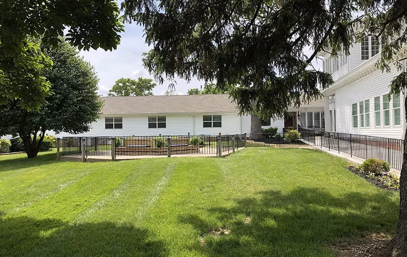 A grassy outdoor area with a fenced garden bed, surrounded by trees and a white building with multiple windows. There is a paved walkway with a railing leading to the building entrance.