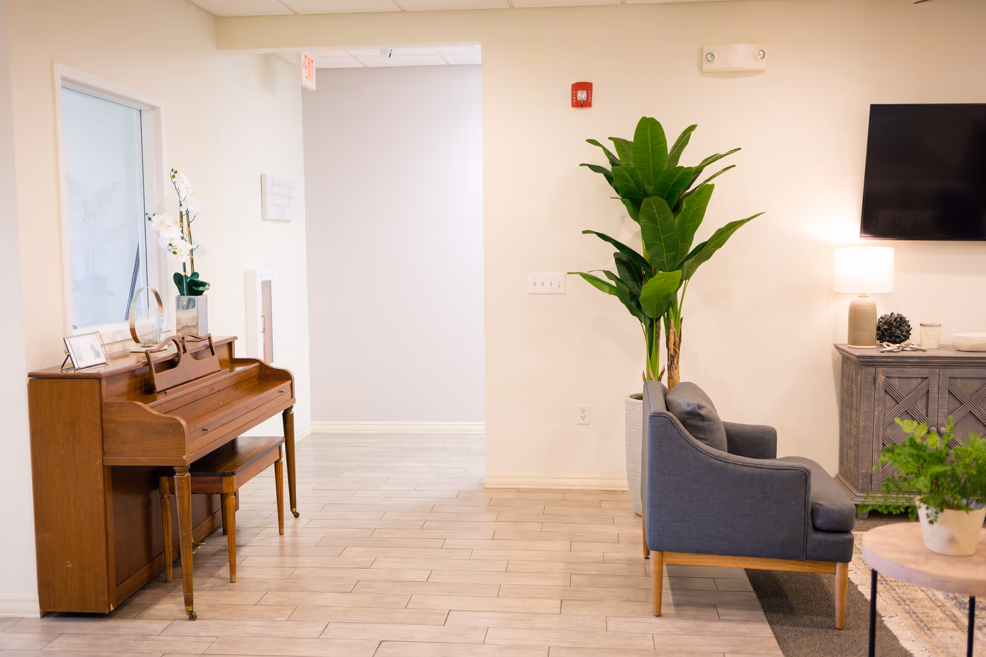 A bright and clean interior space featuring a wooden upright piano with a matching bench on the left, decorated with a white orchid and framed photos. To the right, there is a gray upholstered armchair next to a large green potted plant. A wooden sideboard with a lamp, decorative items, and a wall-mounted flat-screen TV is visible in the background. The floor is light-colored tile, and the walls are painted white.