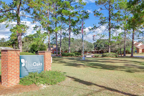 Outdoor view of The Oaks - Bethany assisted living facility with a green sign mounted on brick pillars in the foreground, surrounded by grass and trees under a blue sky with some clouds. The facility buildings and flagpoles are visible in the background.