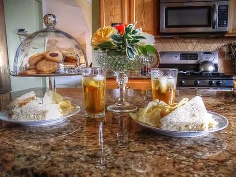 A kitchen countertop with two plates of sandwiches and potato chips, two glasses of iced tea, a glass cake stand with assorted cookies under a glass dome, and a small vase with a bouquet of flowers. In the background, there is a stove, microwave, and wooden cabinets.