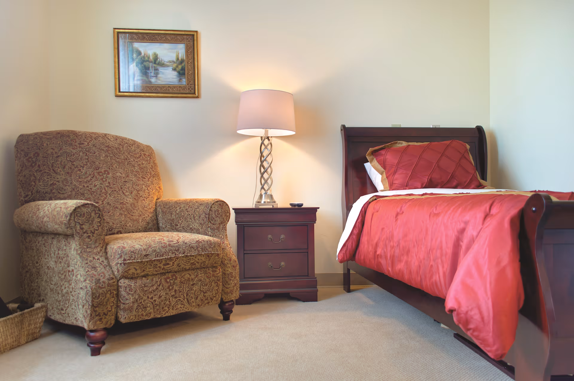 A cozy bedroom featuring a wooden bed with red bedding and a matching pillow, a wooden nightstand with a decorative lamp, a patterned upholstered armchair, and a framed landscape painting on the wall.