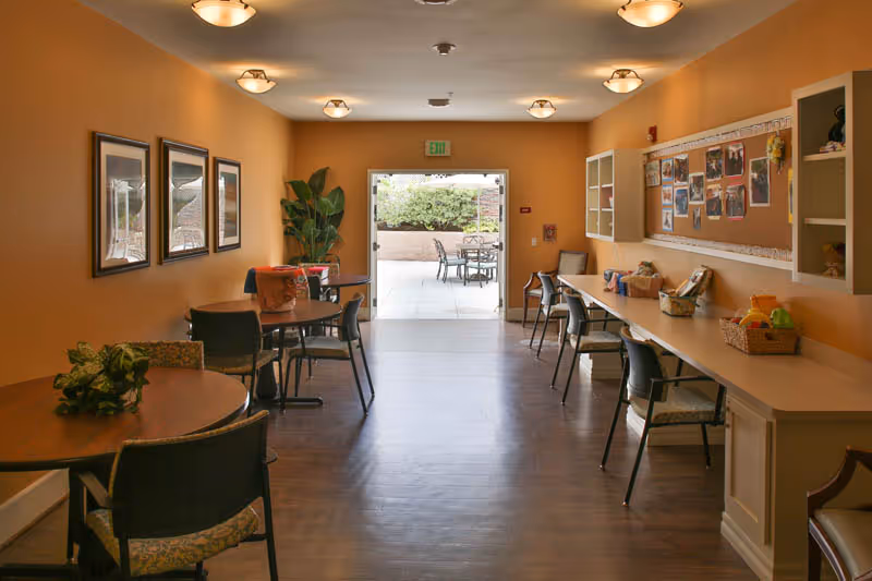 A well-lit interior room with orange walls featuring several tables and chairs arranged for seating. On the right side, there is a long counter with chairs and baskets, along with a bulletin board displaying photos and notes. The room opens to an outdoor patio area with additional tables and chairs visible through a wide open door.