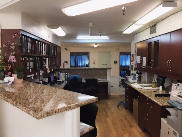 Interior view of a reception or nurse station area in a senior care facility with a granite countertop, office chairs, filing shelves with binders, cabinets, and medical equipment. The room has wooden flooring and fluorescent ceiling lights.
