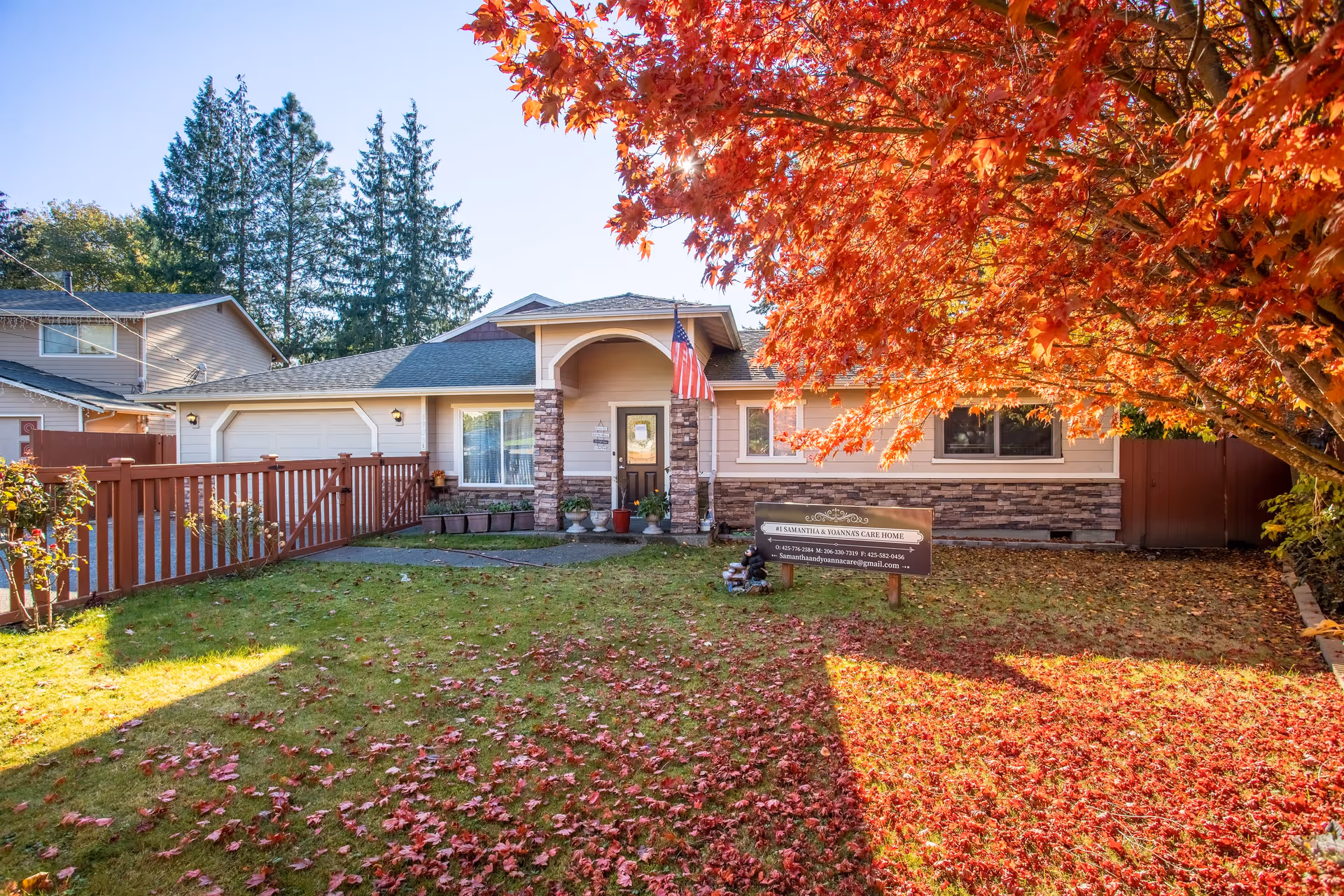 Front exterior view of a single-story care home with a well-maintained lawn covered in autumn leaves. The house features a stone and siding facade, a covered entrance with an American flag, and a wooden fence on the left side. A large tree with bright red fall foliage is on the right side of the image, casting shadows on the lawn. A sign in front of the house reads '#1 Samantha & Yoanna's Care Home' with contact information.
