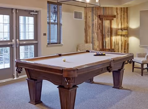 A well-lit room with a wooden pool table in the center, a rack with pool cues mounted on a wooden wall, two upholstered chairs, a floor lamp, and glass doors leading outside.
