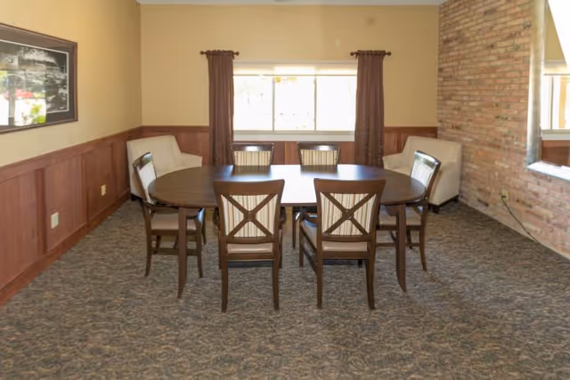 A dining room with a wooden oval table surrounded by six chairs with cushioned seats and backs. Two beige armchairs are placed against the walls on either side of a window with brown curtains. The room has a carpeted floor, wood paneling on the lower half of the walls, and a brick accent wall with a large mirror.