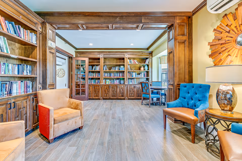 A cozy senior living common area with wooden bookshelves filled with books, two beige armchairs, a blue cushioned chair, a round side table with a lamp, and a decorative wooden sunburst mirror on the wall. The room has wood flooring and warm wood paneling around the bookshelves and door frames.