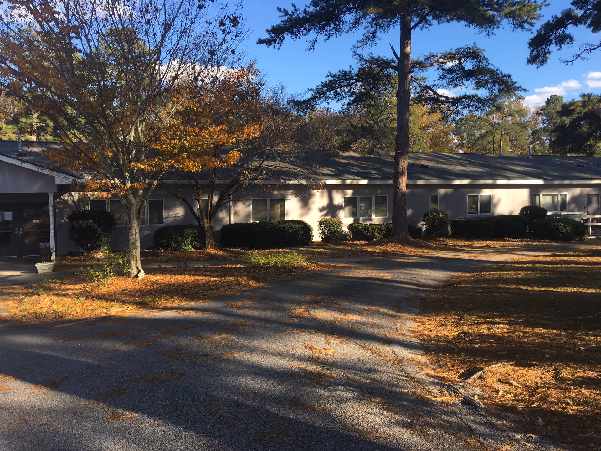 Exterior view of a single-story healthcare facility building surrounded by trees with autumn foliage and bushes. A paved driveway leads to the entrance on the left side of the building under a clear blue sky.