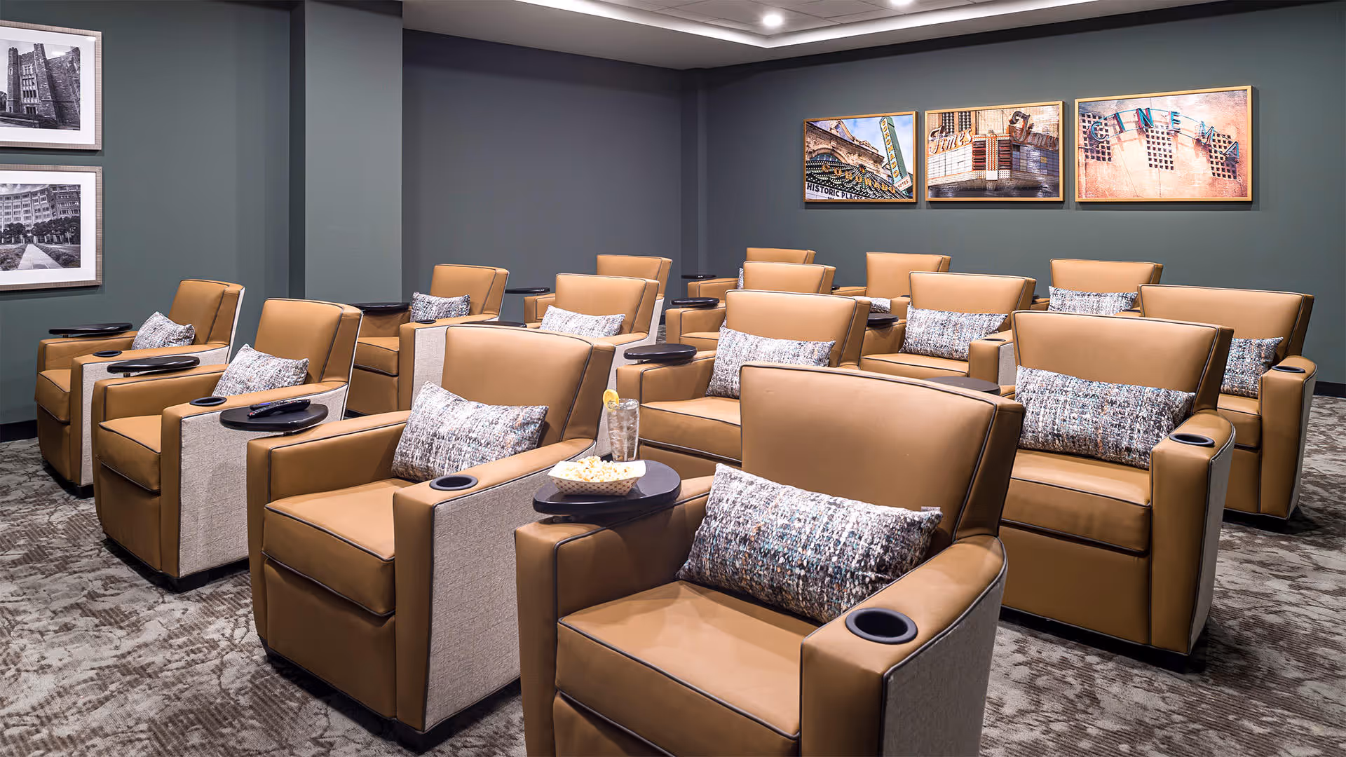 A cozy theater-style room with multiple rows of tan leather recliner chairs, each equipped with a side table and a decorative pillow. The walls are painted a muted green and adorned with framed artwork. The carpet has a patterned design, and the room is softly lit with recessed ceiling lights.