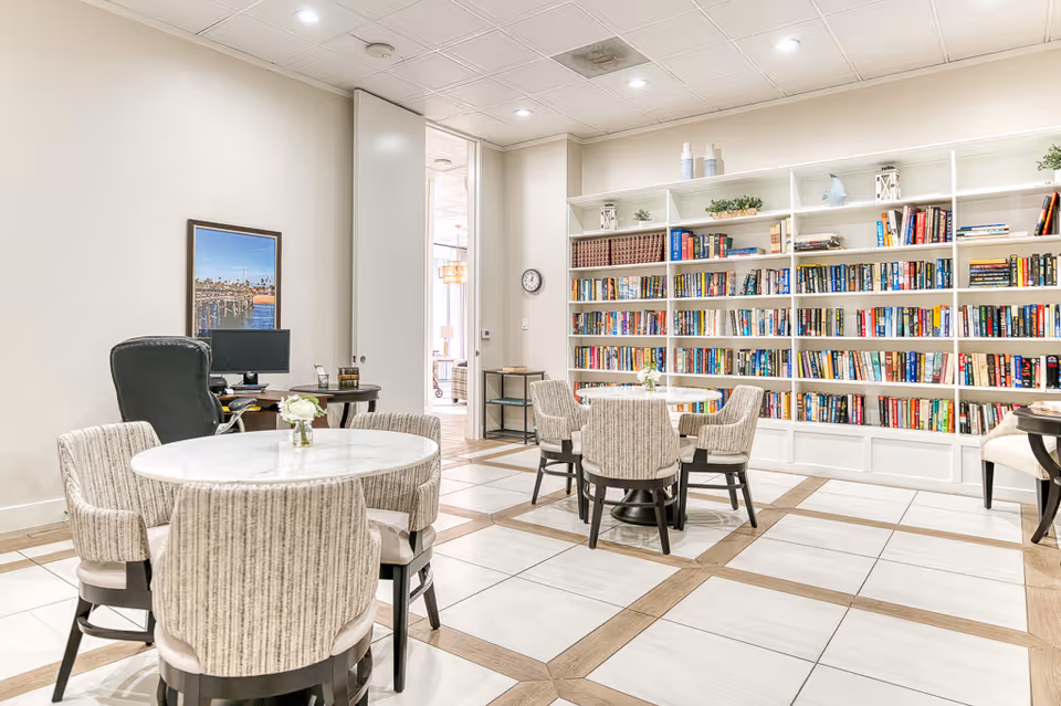 A bright and spacious room with white tiled floors and wooden accents, featuring round tables with cushioned chairs arranged for seating. One wall is lined with a large white bookshelf filled with books and decorative items. A desk with a computer and a black office chair is positioned against another wall, and a framed picture hangs above it. The room has a clean, modern look with recessed lighting in the ceiling.