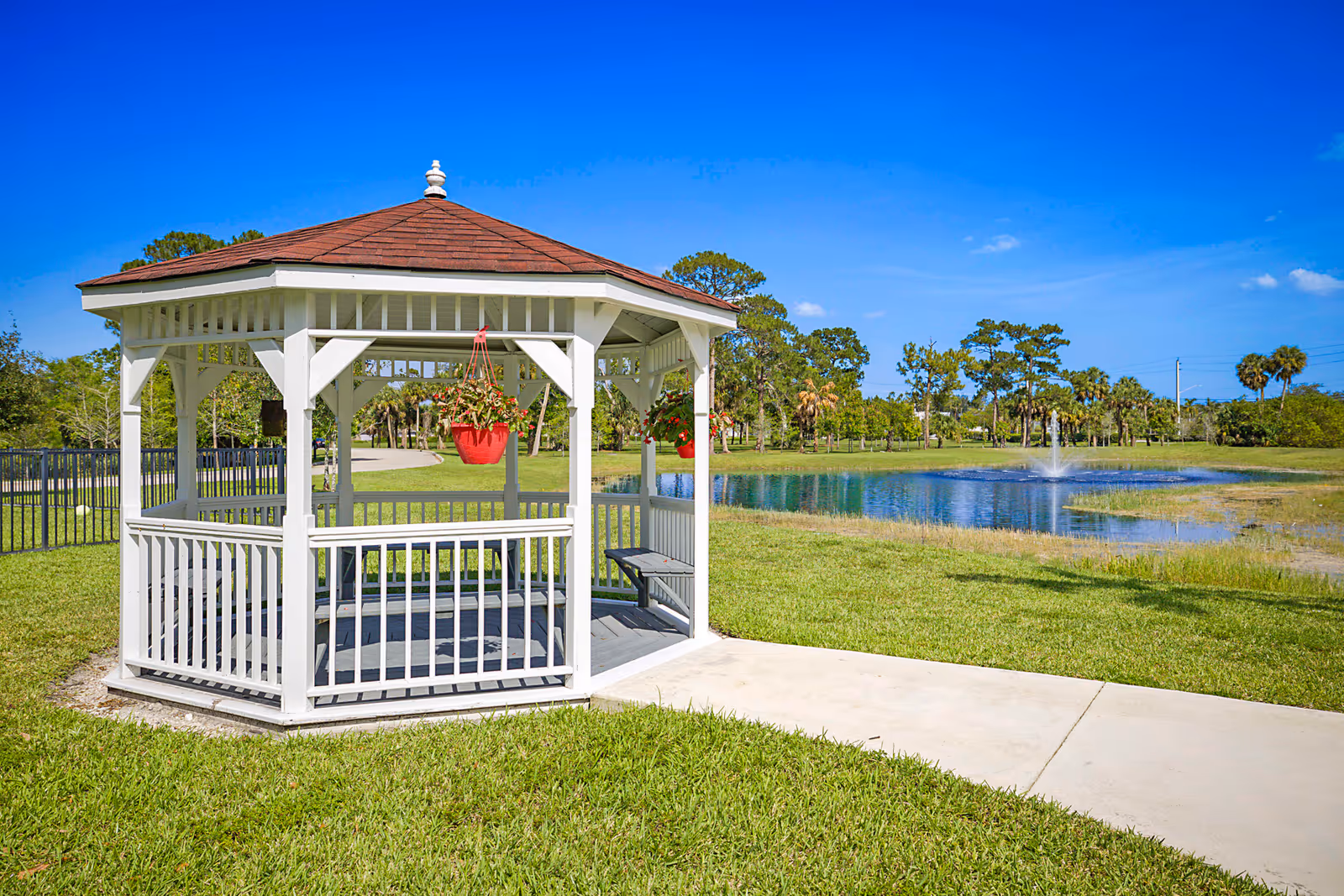 A white wooden gazebo with hanging red flower pots beside a pond and fountain on a sunny day.