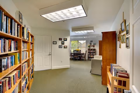 Interior view of a senior living facility room with bookshelves filled with books on the left and right sides. The room has carpeted flooring, white walls with framed pictures, and fluorescent ceiling lights. In the background, there is a seating area with a table and chairs near a window letting in natural light.