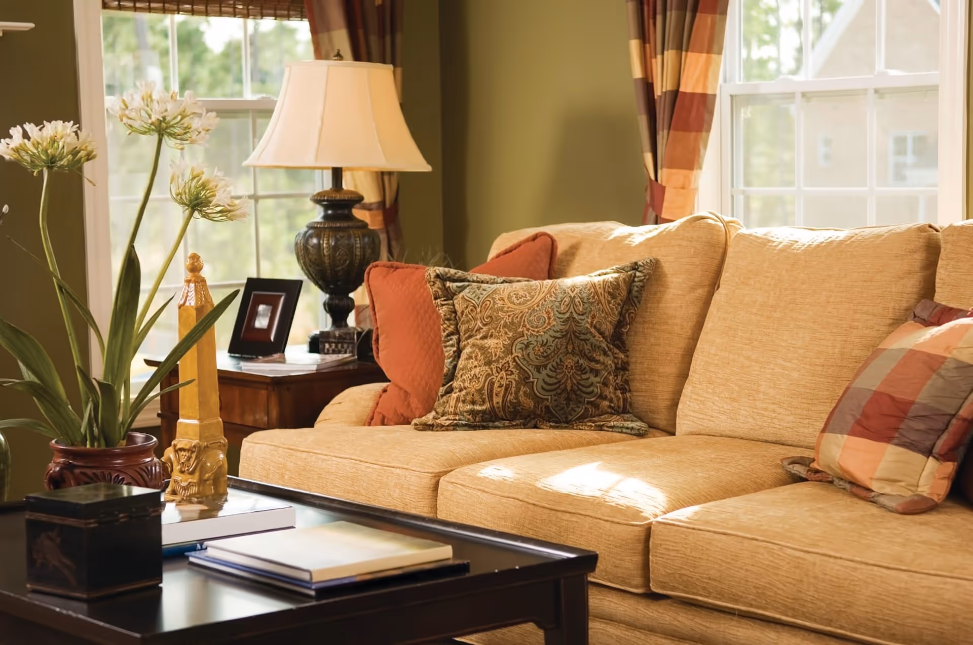 A cozy living room with a beige sofa adorned with patterned and solid throw pillows. A dark wooden coffee table in front holds books and decorative items. Behind the sofa, there is a side table with a classic lamp and a small framed photo. Two large windows with plaid curtains let in natural light, and a potted plant with white flowers adds a touch of greenery.