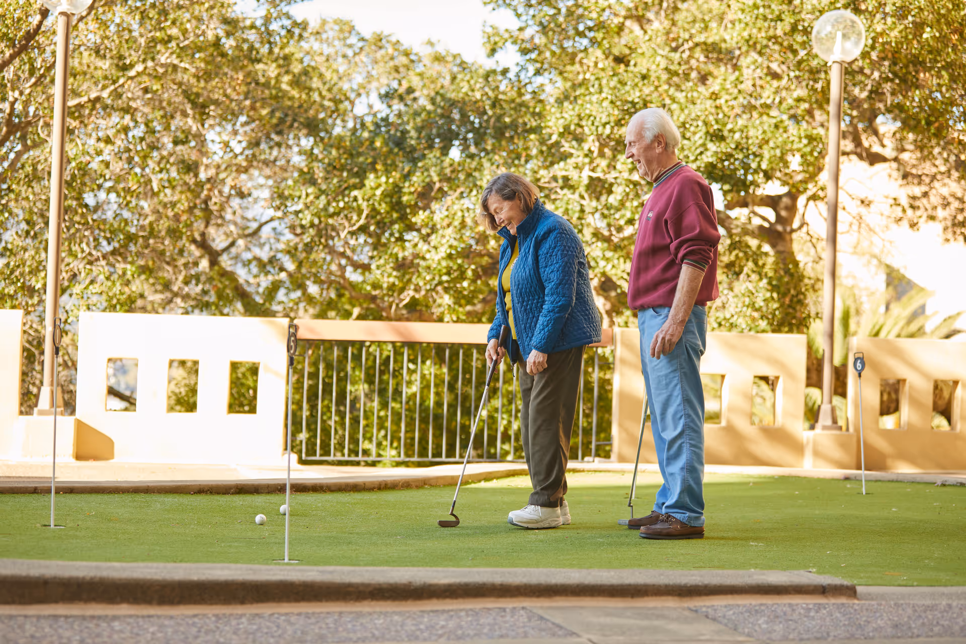 An elderly woman and man playing mini golf on a putting green outdoors, surrounded by trees and a beige wall with square cutouts. The woman is putting while the man watches and smiles.