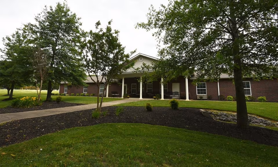 Single-story brick building with a columned front porch, walkway, lawns and trees in the foreground.
