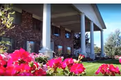 Exterior view of a senior living facility with large white columns supporting a covered porch. The building has a brick facade and multiple windows. In the foreground, there are vibrant pink flowers and green grass under a clear blue sky.