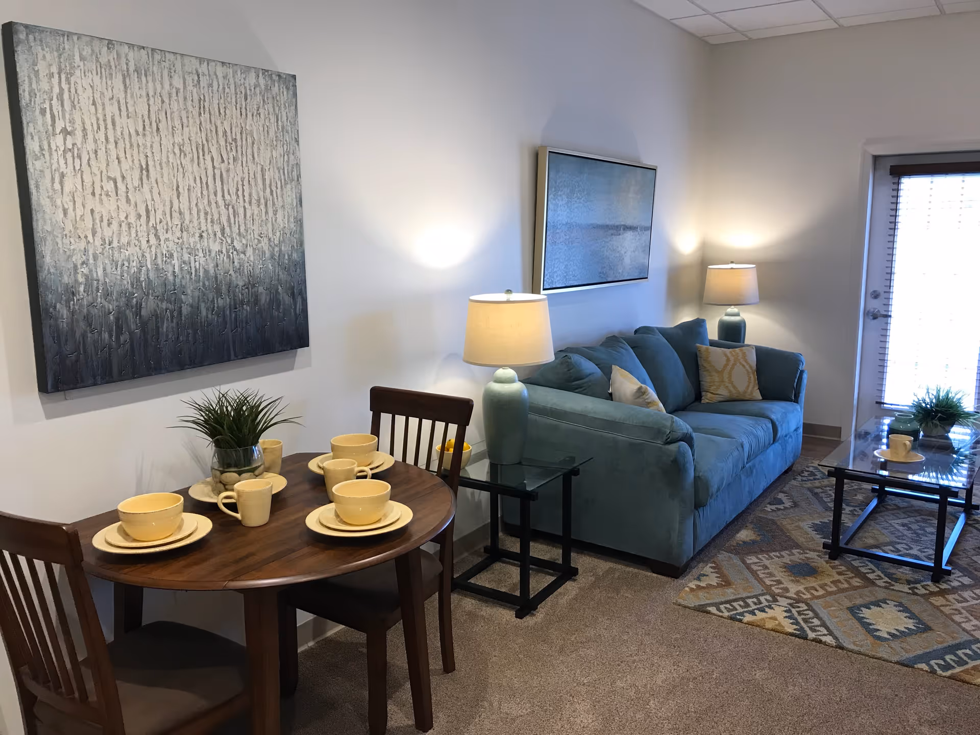A cozy living area featuring a teal sofa with decorative pillows, two table lamps on glass side tables, a wooden round dining table set with yellow dishes and cups, a patterned rug, and two abstract paintings on the walls. A glass door with blinds is visible in the background.