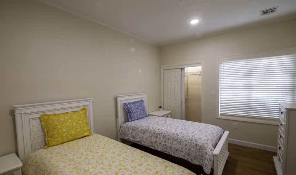 A bedroom with two single beds, each with a white wooden headboard. One bed has yellow floral bedding and a matching pillow, while the other has blue floral bedding and a matching pillow. There is a small white nightstand between the beds with a small plant on it. A closet with sliding doors is visible in the background, along with a window with white blinds. The room has light-colored walls and wooden flooring.