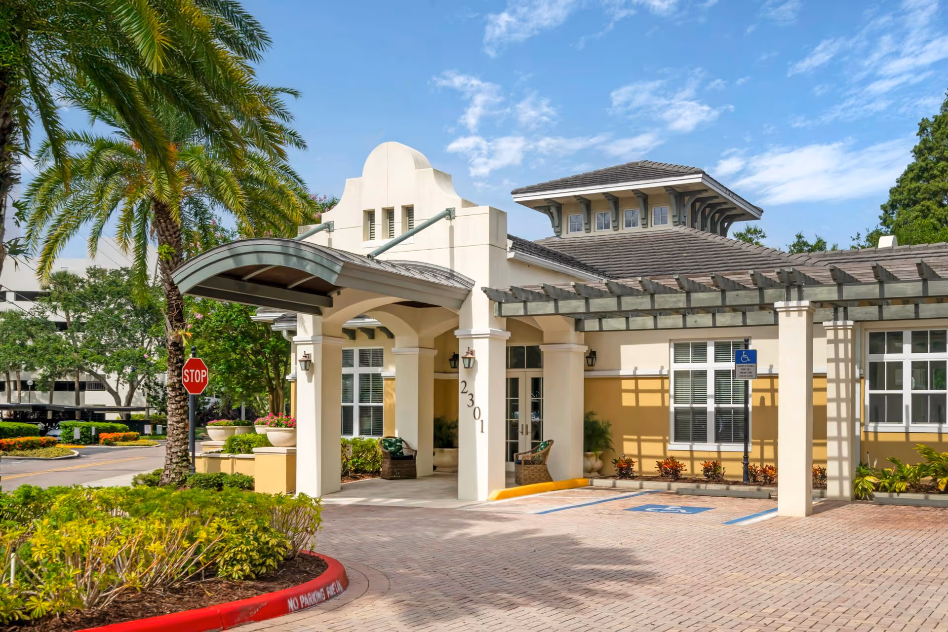 Entrance of The Estate at Hyde Park facility featuring a covered drop-off area with columns, a stop sign, palm trees, and a clear blue sky.