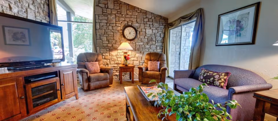Cozy living room with two recliners and a sofa facing a TV, a stone accent wall with a clock, and a coffee table with a plant.