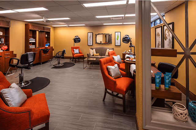 Interior view of a salon area in a senior living facility with orange upholstered chairs, salon stations with mirrors and hair dryers, a small seating area with a sofa and chairs, warm yellow walls, and framed artwork.