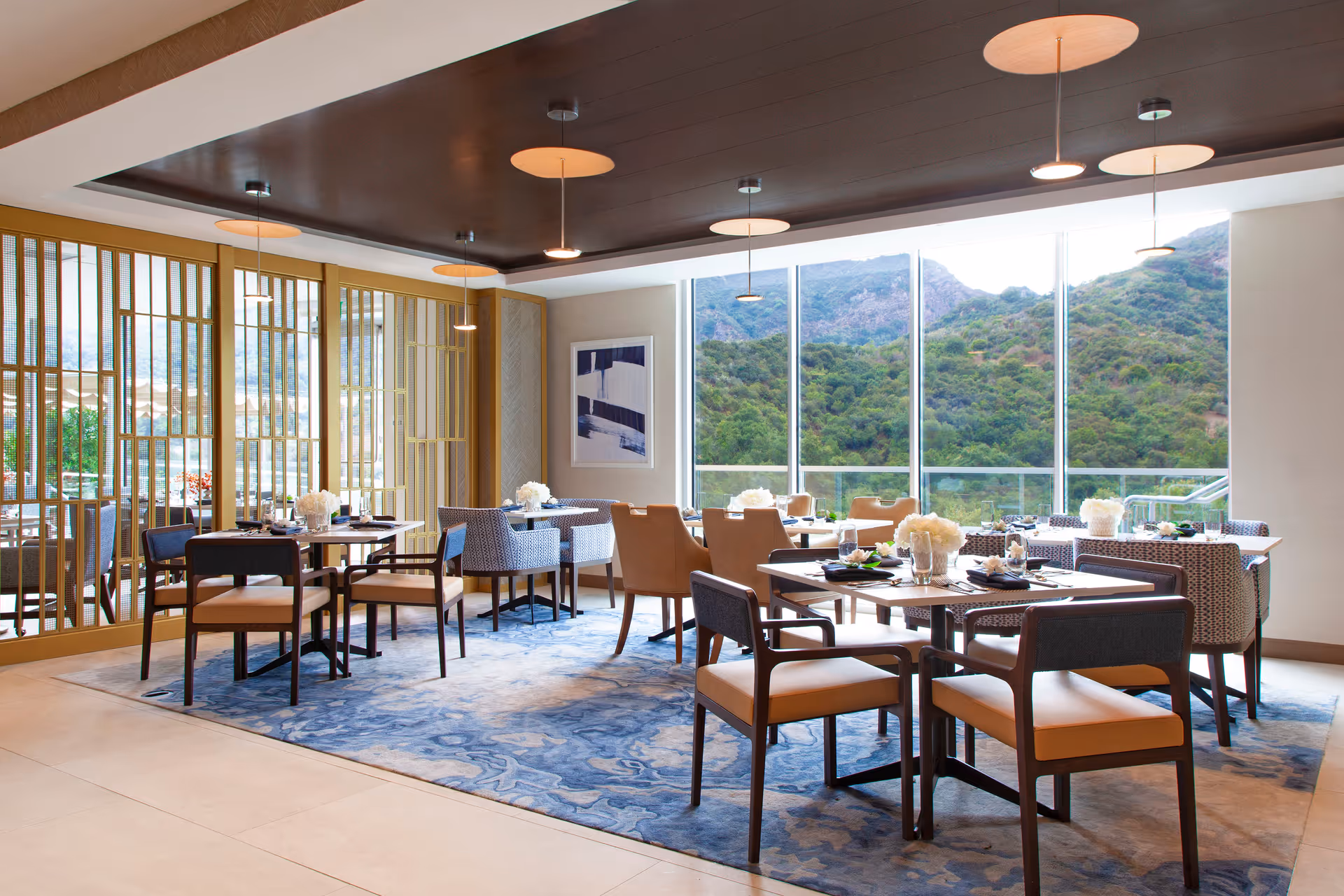 A bright and modern dining room with several tables and chairs arranged on a blue patterned rug. Large floor-to-ceiling windows offer a scenic view of green hills and mountains outside. The room features pendant lights hanging from a dark ceiling panel and decorative wooden partitions on one side.