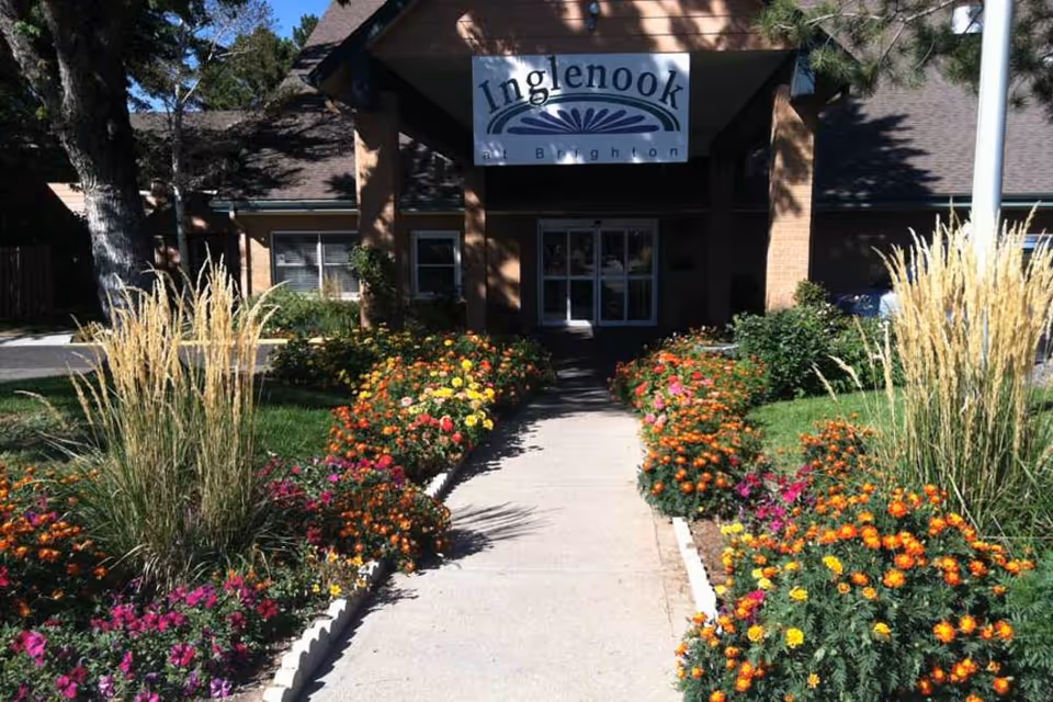 Pathway lined with colorful flowers and tall grasses leading to the entrance of a building with a sign that reads 'Inglenook at Brighton'. The building has a brick exterior and a peaked roof.