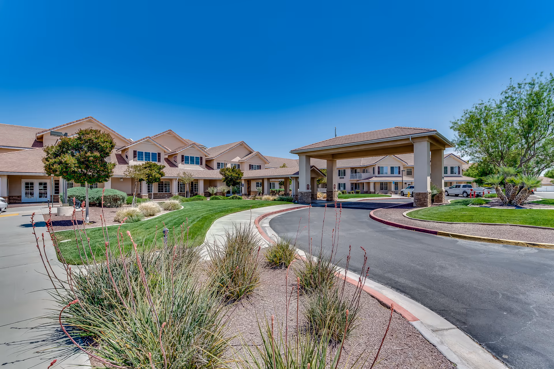 Exterior view of a senior living facility with a covered driveway entrance, landscaped greenery, trees, and a clear blue sky.