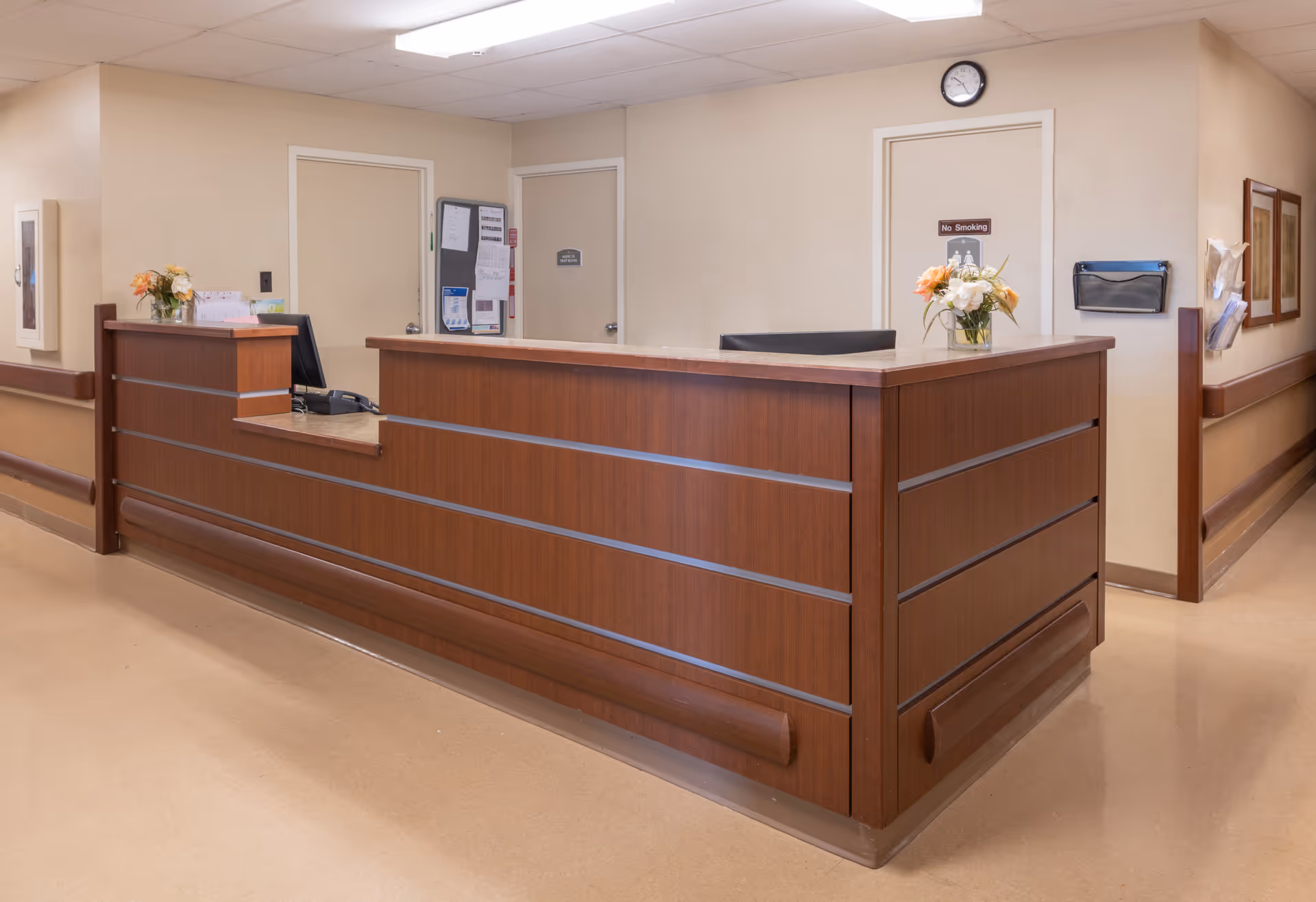 Reception desk area in a nursing and rehabilitation facility with a wooden counter, two vases of flowers, a computer monitor, and doors in the background, one with a 'No Smoking' sign and a clock above it.