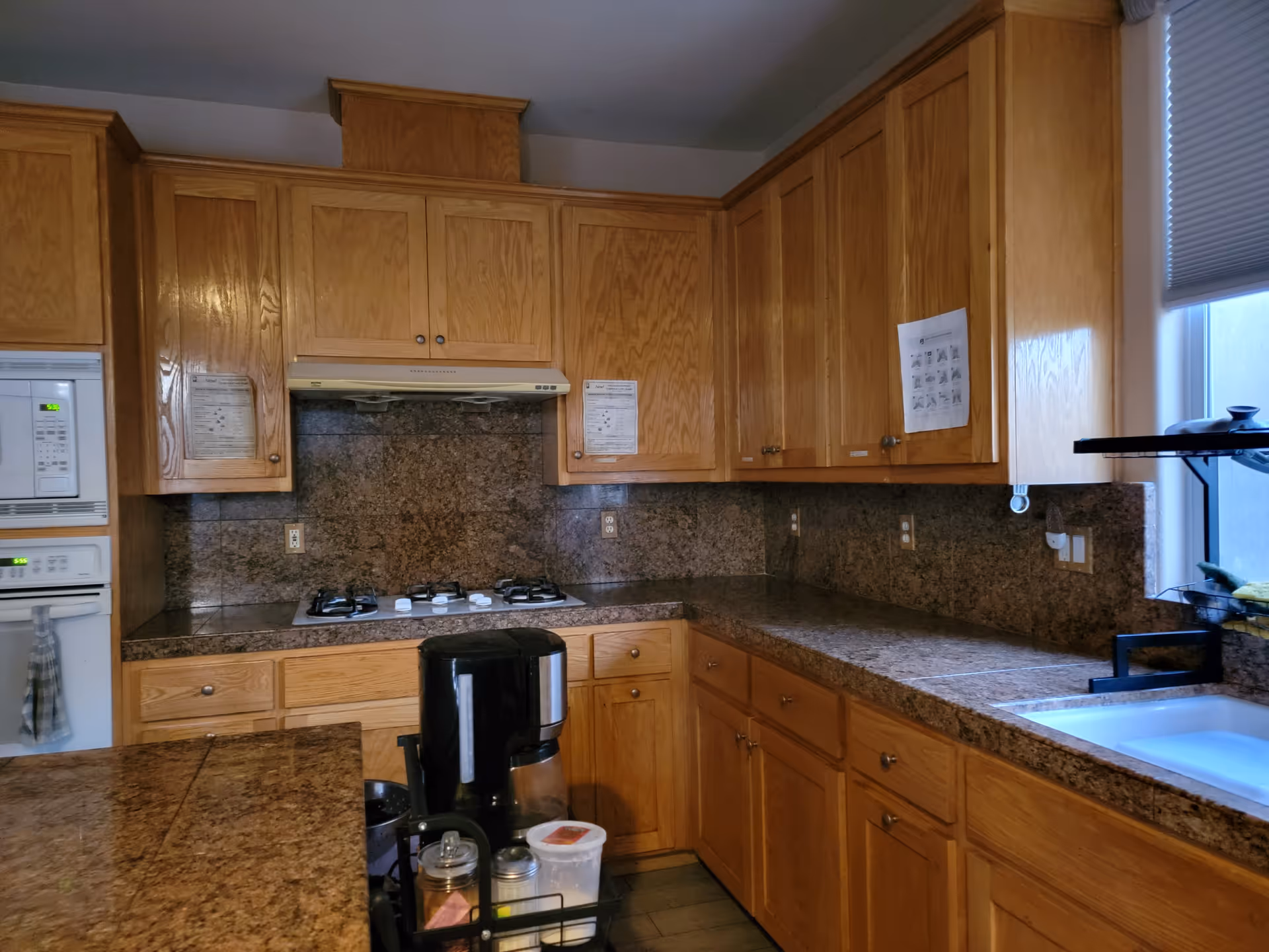 A kitchen with wooden cabinets and granite countertops. The kitchen features a built-in microwave, oven, and a gas stove with a range hood. There is a coffee maker and various containers on the counter. A window with a blind is visible above the sink.
