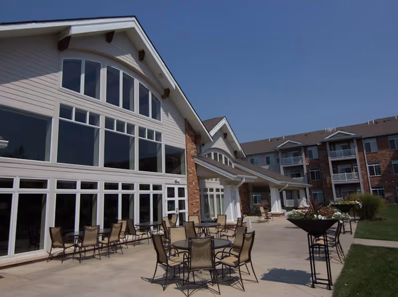 Outdoor patio area of a senior living facility with multiple tables and chairs arranged on a concrete surface. The building features large windows and a peaked roof, with a well-maintained lawn and flower planters nearby under a clear blue sky.