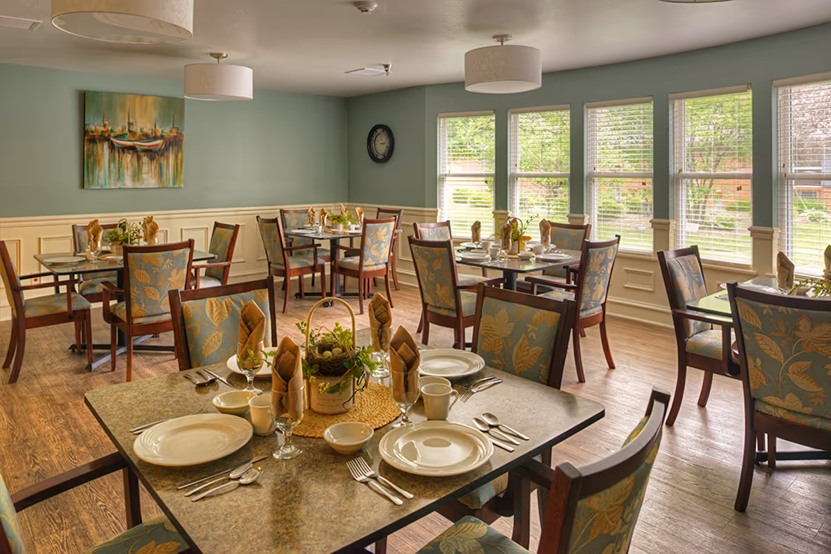 A dining room with multiple tables set for meals, each table has plates, cups, utensils, and napkins. The chairs have a floral pattern upholstery. Large windows with blinds allow natural light to brighten the room. A painting and a clock hang on the walls.