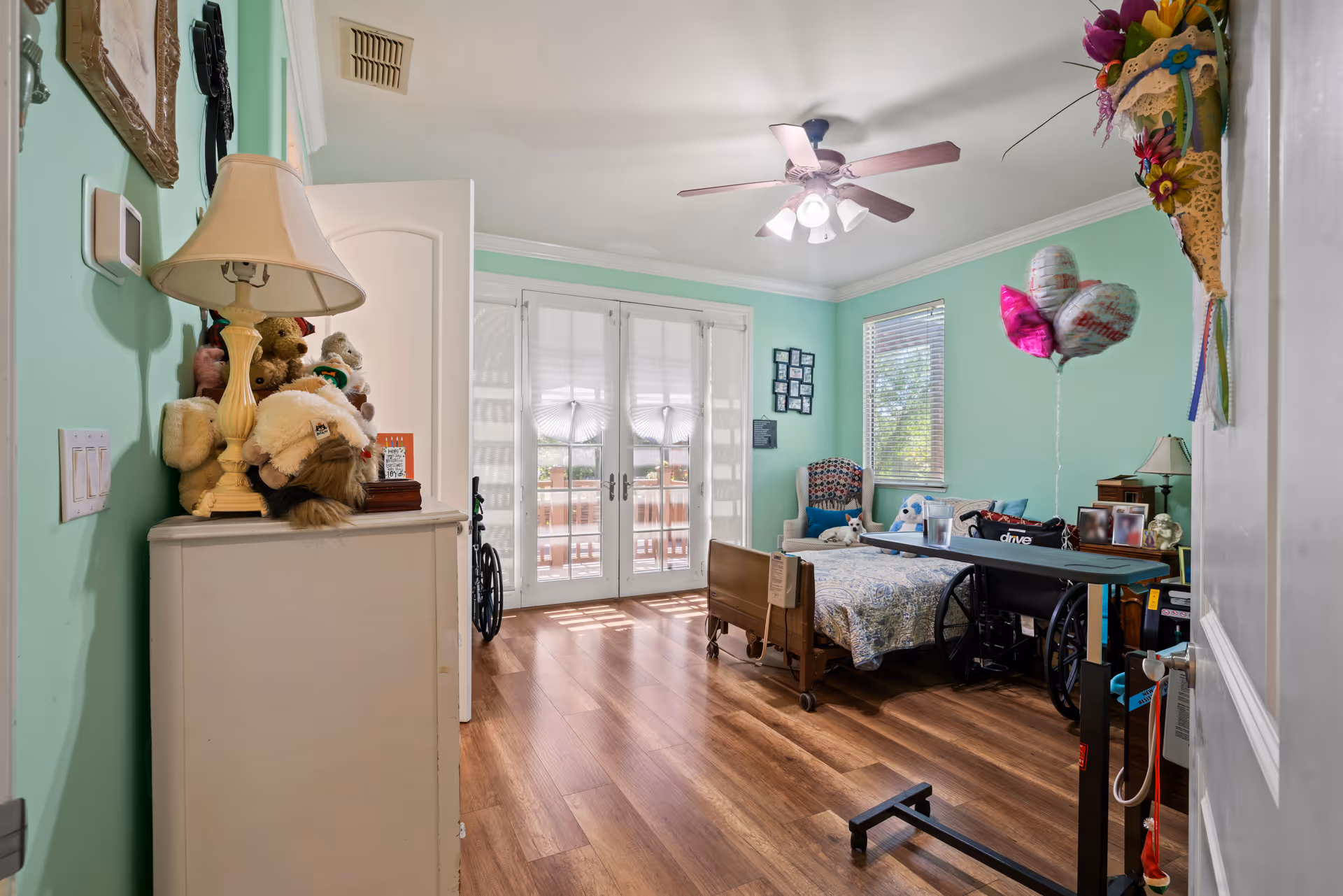 A bright bedroom with mint green walls and wood flooring. The room features a hospital-style bed with a patterned bedspread, a wheelchair, a small dog sitting on a chair, and a ceiling fan with lights. There are French doors with white blinds leading outside, a dresser with a lamp and stuffed animals, and birthday balloons near the bed. A floral wall decoration hangs on the right side of the door.