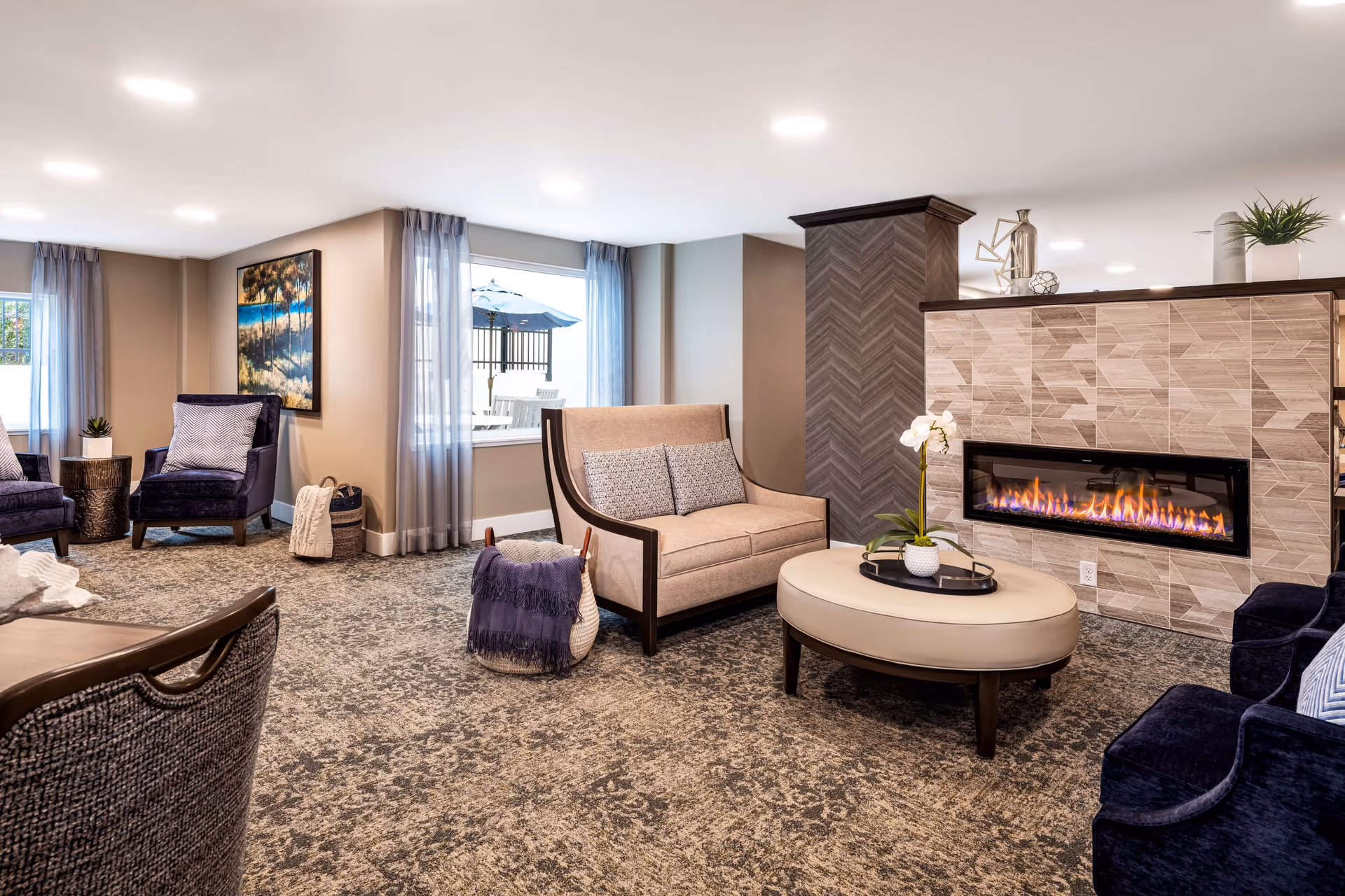 Bright, furnished lounge with armchairs, a sofa, round ottoman, and a modern fireplace.