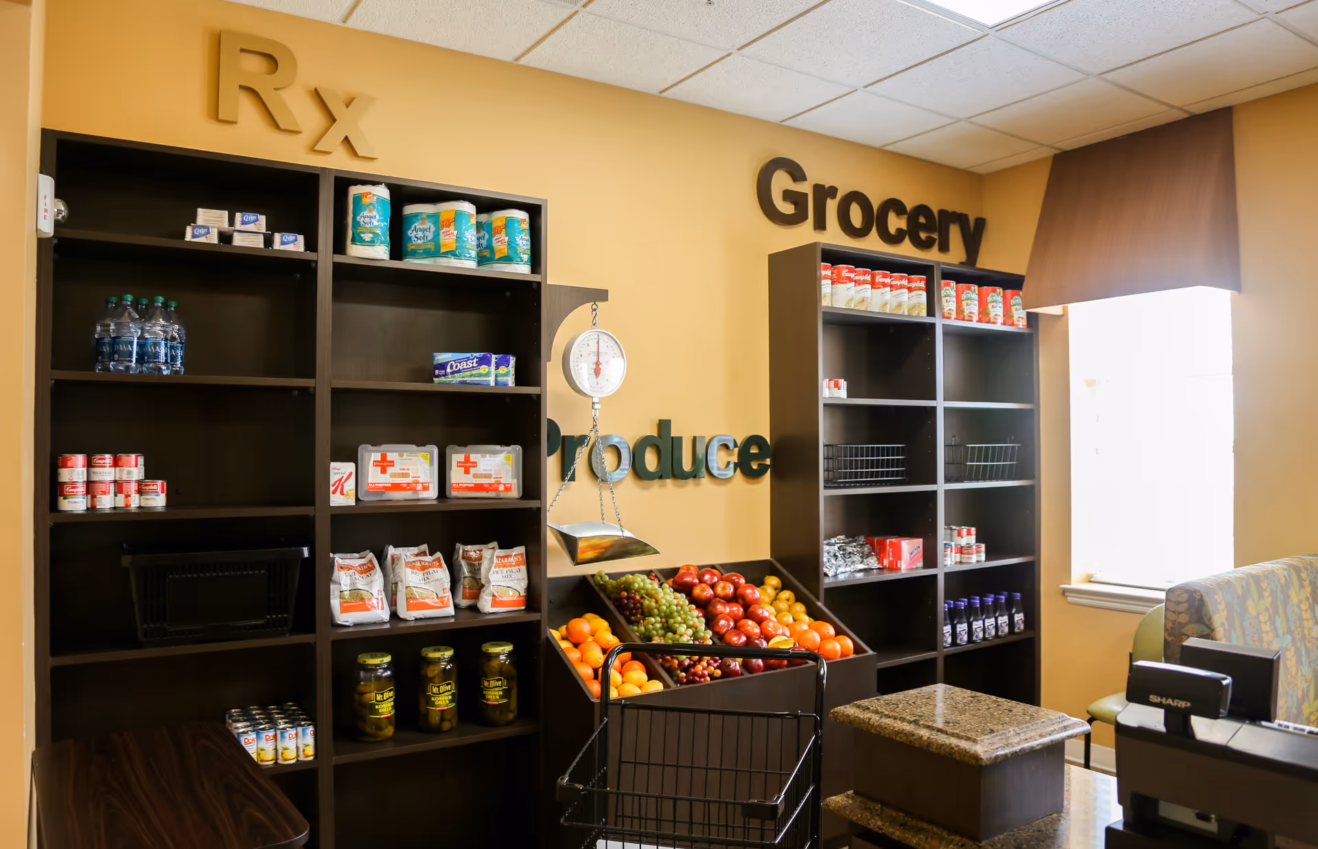 Interior view of a small grocery and produce area in a senior living facility. Shelves are stocked with canned goods, bottled water, paper towels, and other grocery items. A produce display holds fresh fruits like apples, grapes, and oranges. A hanging scale is visible above the produce section. The walls have signs reading 'Rx', 'Produce', and 'Grocery'. A window with a brown valance lets in natural light.