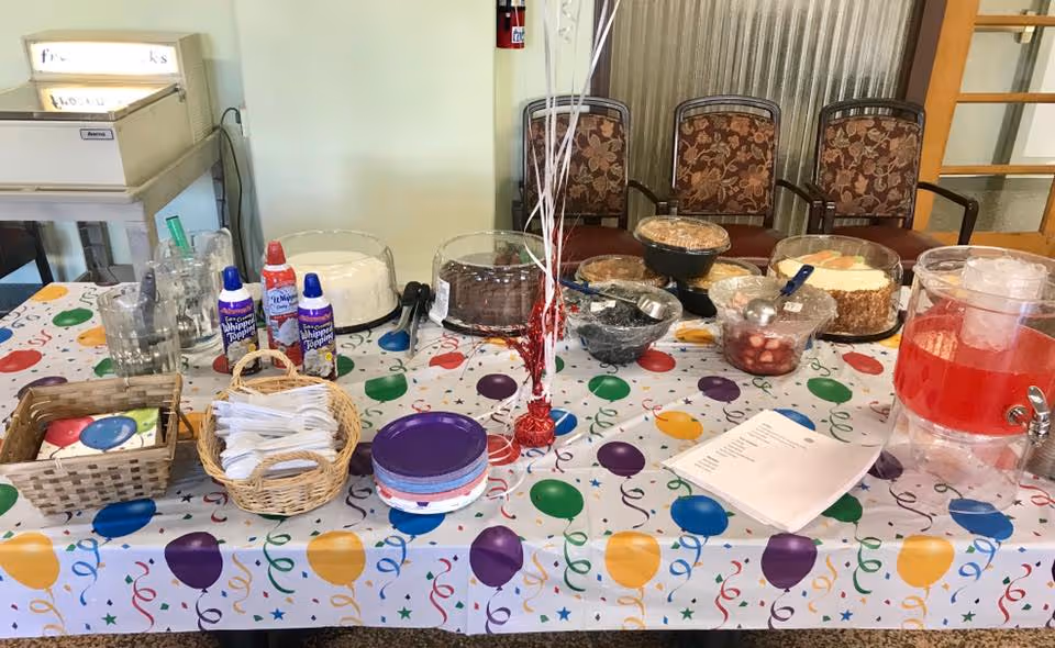 A decorated buffet table with plates, utensils, cakes, bowls of fruit and a red beverage dispenser set up in a communal dining area.