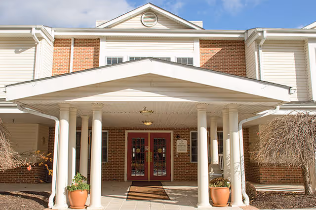 The covered brick entrance of a senior living building with white columns, red double doors, and potted plants.