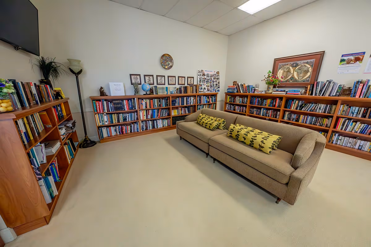 A cozy library or reading room with beige carpeting, a brown couch with patterned cushions, and multiple wooden bookshelves filled with books. The walls are decorated with framed pictures, a globe, and a floor lamp is placed in the corner. A flat-screen TV is mounted on the wall.