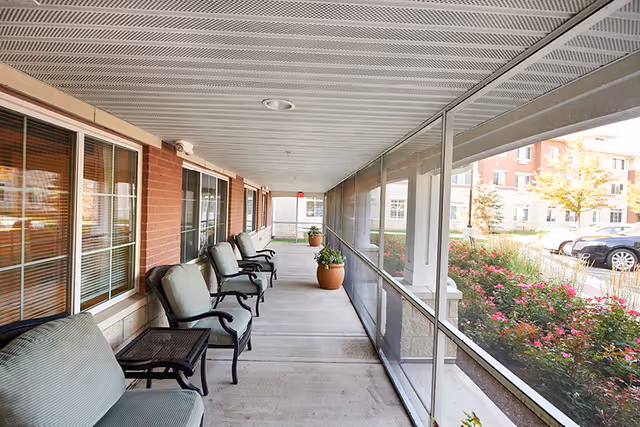 Covered outdoor patio area with cushioned chairs and small tables lined up along a brick wall with windows on one side and a screened enclosure on the other. Potted plants are placed along the walkway, and outside the screened area, there are bushes with pink flowers and parked cars visible.