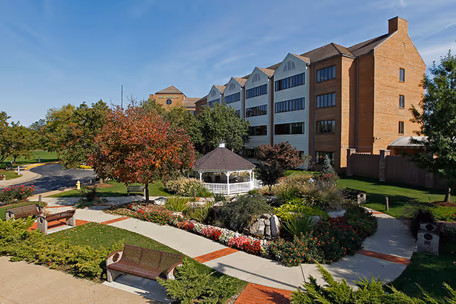 A large multi-story brick and white building with many windows, surrounded by a well-maintained garden featuring a white gazebo, colorful flowers, shrubs, and trees with autumn foliage. There are paved walkways and benches in the garden area under a clear blue sky.