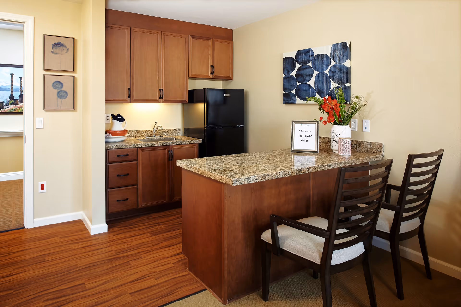 A small kitchen area with wooden cabinets, a black refrigerator, and a granite countertop island with two wooden chairs. The countertop has a vase with flowers and a small sign indicating a 1 bedroom floor plan B2 with 607 square feet. There is a piece of abstract blue and white artwork on the wall and two framed floral prints on the adjacent wall near a doorway.