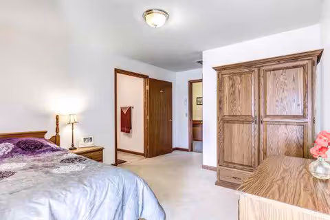 A bedroom with a bed covered in a purple and gray patterned comforter, a wooden nightstand with a lamp and a clock, a wooden wardrobe, and a wooden dresser with a vase of pink flowers. The room has white walls, beige carpet, and two open doorways leading to other rooms.