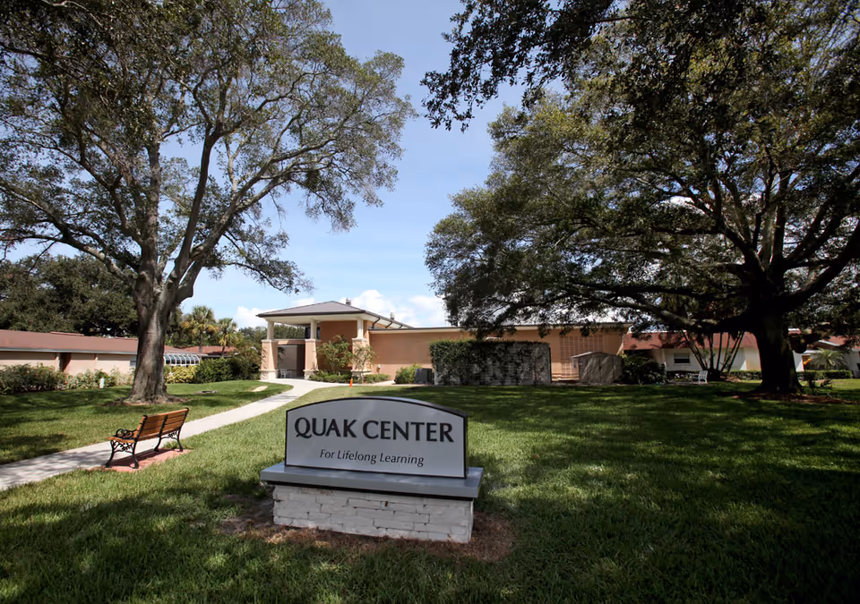 Front lawn and entrance of the Quak Center building with a sign reading "QUAK CENTER For Lifelong Learning", a walkway, bench, and large trees.